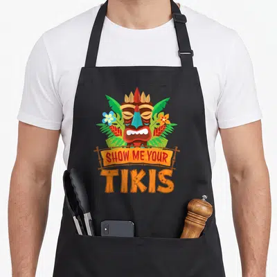 Tikis hawaiian apron - Cartoon tiki mask with green leaves and orange banner reading SHOW ME YOUR TIKIS — shown on a black cotton canvas apron against a white background