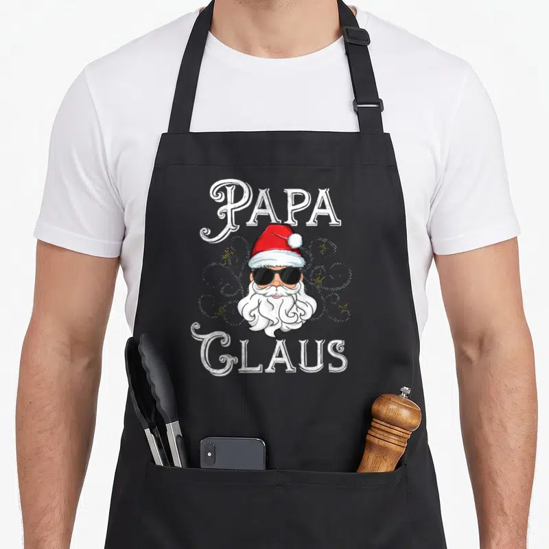 Claus family christmas aprons - Cartoon Santa head with sunglasses between PAPA and GLAUS text in white red black — shown on a black cotton canvas apron against a white background