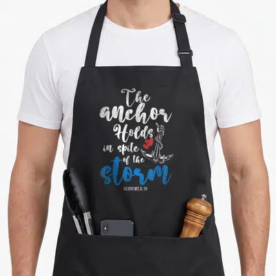 Holy anchor aprons - White and blue text with anchor and two red hearts — shown on a black cotton canvas apron against a white background
