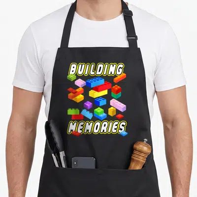 Memory building apron - BUILDING MEMORIES text surrounding scattered red, blue, green, pink, purple, yellow, and white — shown on a black cotton canvas apron against a white background