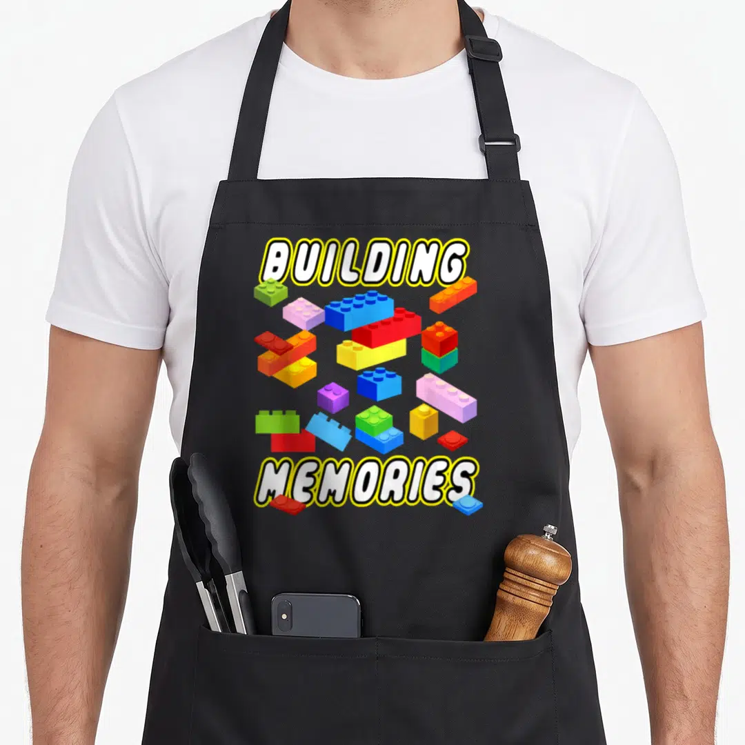 Memory building apron - BUILDING MEMORIES text surrounding scattered red, blue, green, pink, purple, yellow, and white — shown on a black cotton canvas apron against a white background