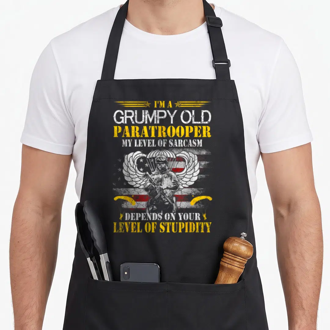 Grumpy paratrooper apron - Soldier with rifle in front of distressed flag and wings with sarcastic text — shown on a black cotton canvas apron against a white background