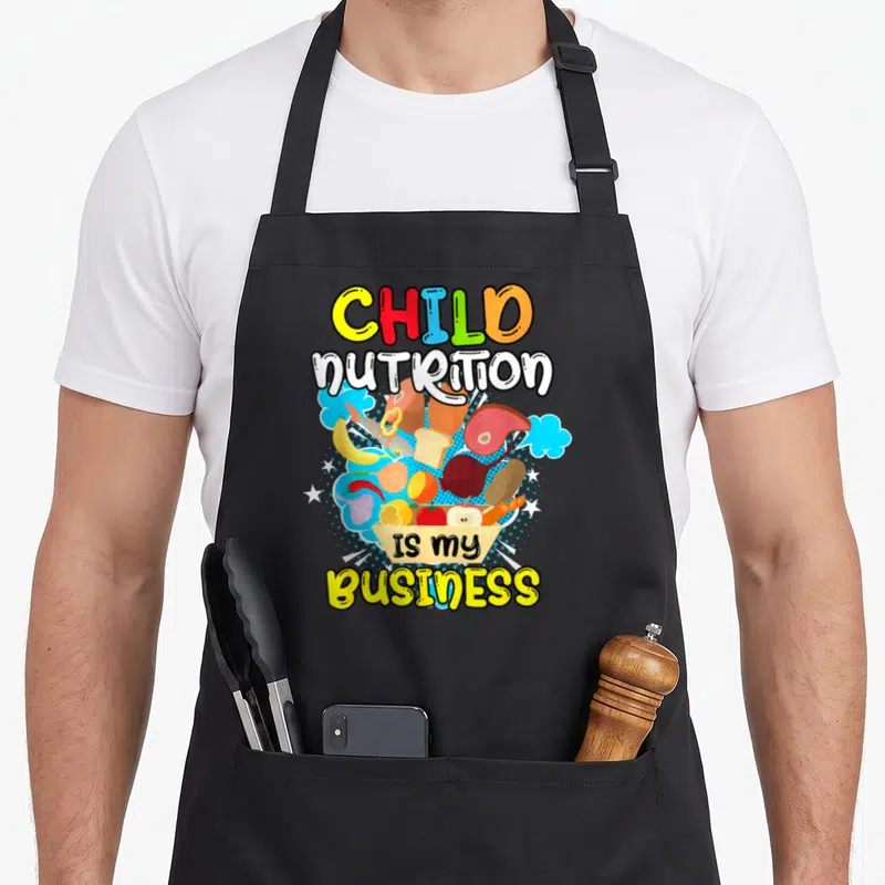 Business cafeteria aprons - Colorful cartoon food cluster with blue clouds and text reading Child Nutrition Is My — shown on a black cotton canvas apron against a white background