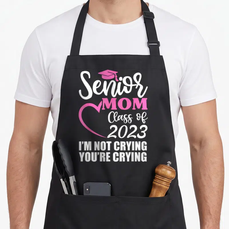 Senior mom class apron - Pink grad cap and heart outline with Senior MOM Class of 2023 and I'm not crying you're crying — shown on a black cotton canvas apron against a white background