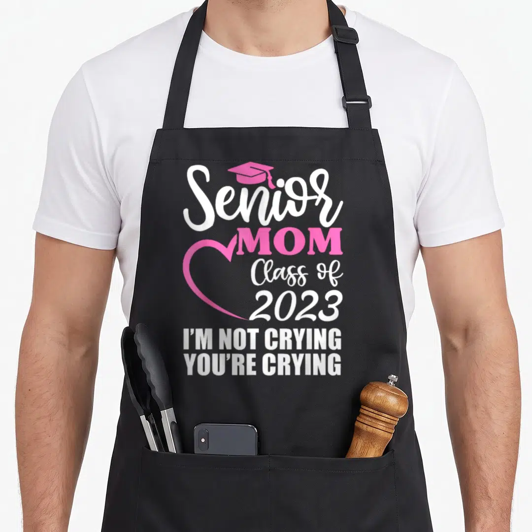 Senior mom class apron - Pink grad cap and heart outline with Senior MOM Class of 2023 and I'm not crying you're crying — shown on a black cotton canvas apron against a white background
