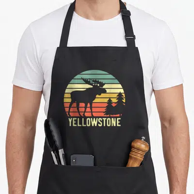 Wyoming travel life apron - Retro moose silhouette and two pine trees against striped sunset with YELLOWSTONE text — shown on a black cotton canvas apron against a white background