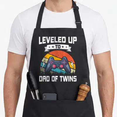 Leveled gaming apron - Retro controller over striped sun with LEVELED UP TO DAD OF TWINS text — shown on a black cotton canvas apron against a white background