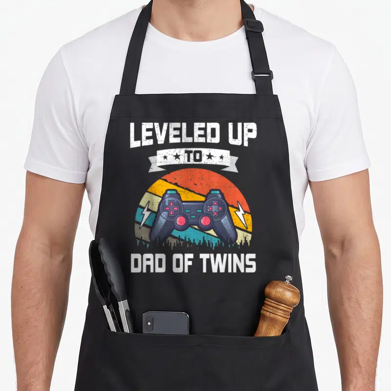 Leveled gaming apron - Retro controller over striped sun with LEVELED UP TO DAD OF TWINS text — shown on a black cotton canvas apron against a white background