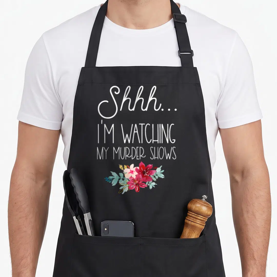 Shhh true crime apron - Stacked white text reading Shhh... I'm Watching My Murder Shows above red flowers and teal — shown on a black cotton canvas apron against a white background
