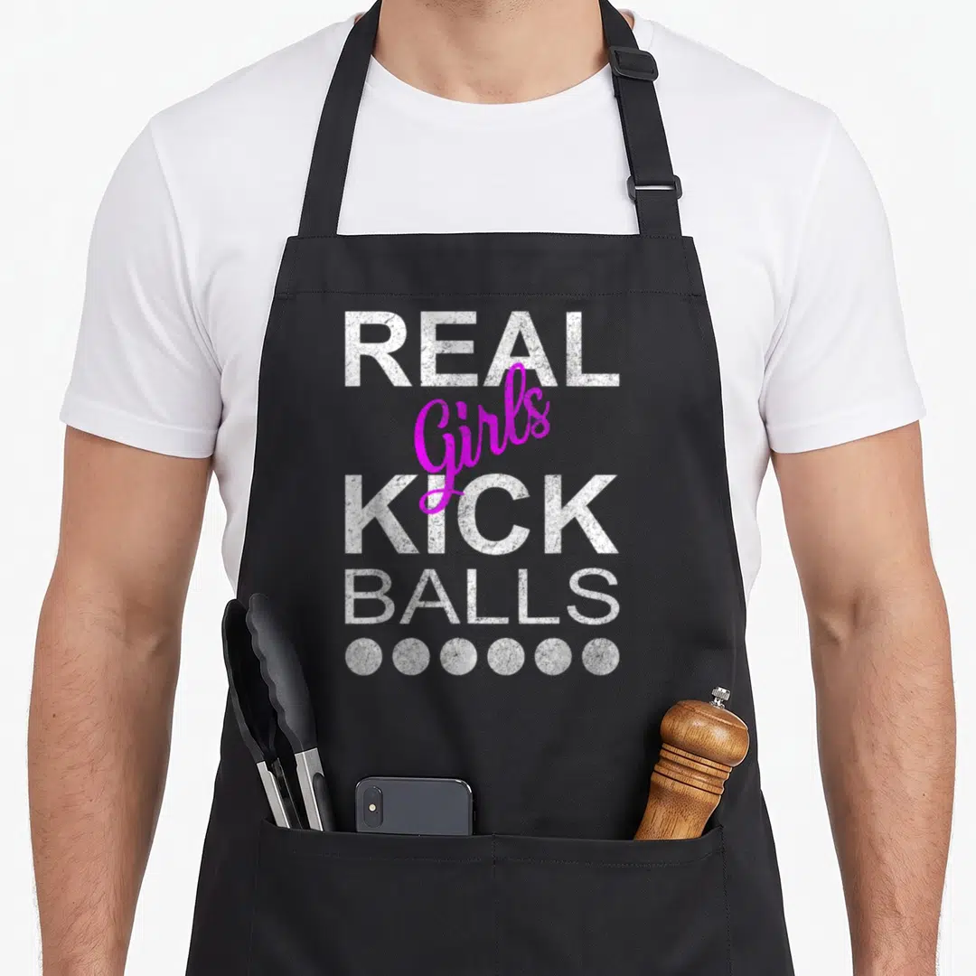 Kick ball kickball apron - Stacked REAL Girls KICK BALLS text with six white circles below in white and magenta — shown on a black cotton canvas apron against a white background