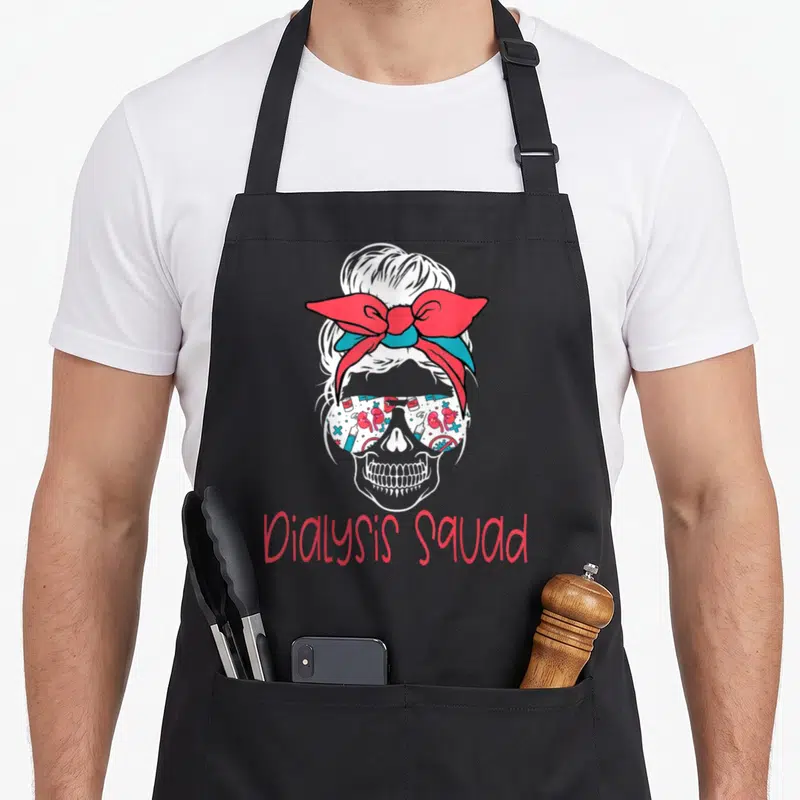 Hemodialysis nurse apron - White skull with red teal bow, medical icon sunglasses, red Dialysis Squad text below — shown on a black cotton canvas apron against a white background