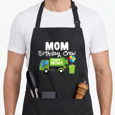 Garbage crew aprons - MOM Birthday Crew text above green truck with HAPPY BIRTHDAY! and balloons in bin — shown on a black cotton canvas apron against a white background