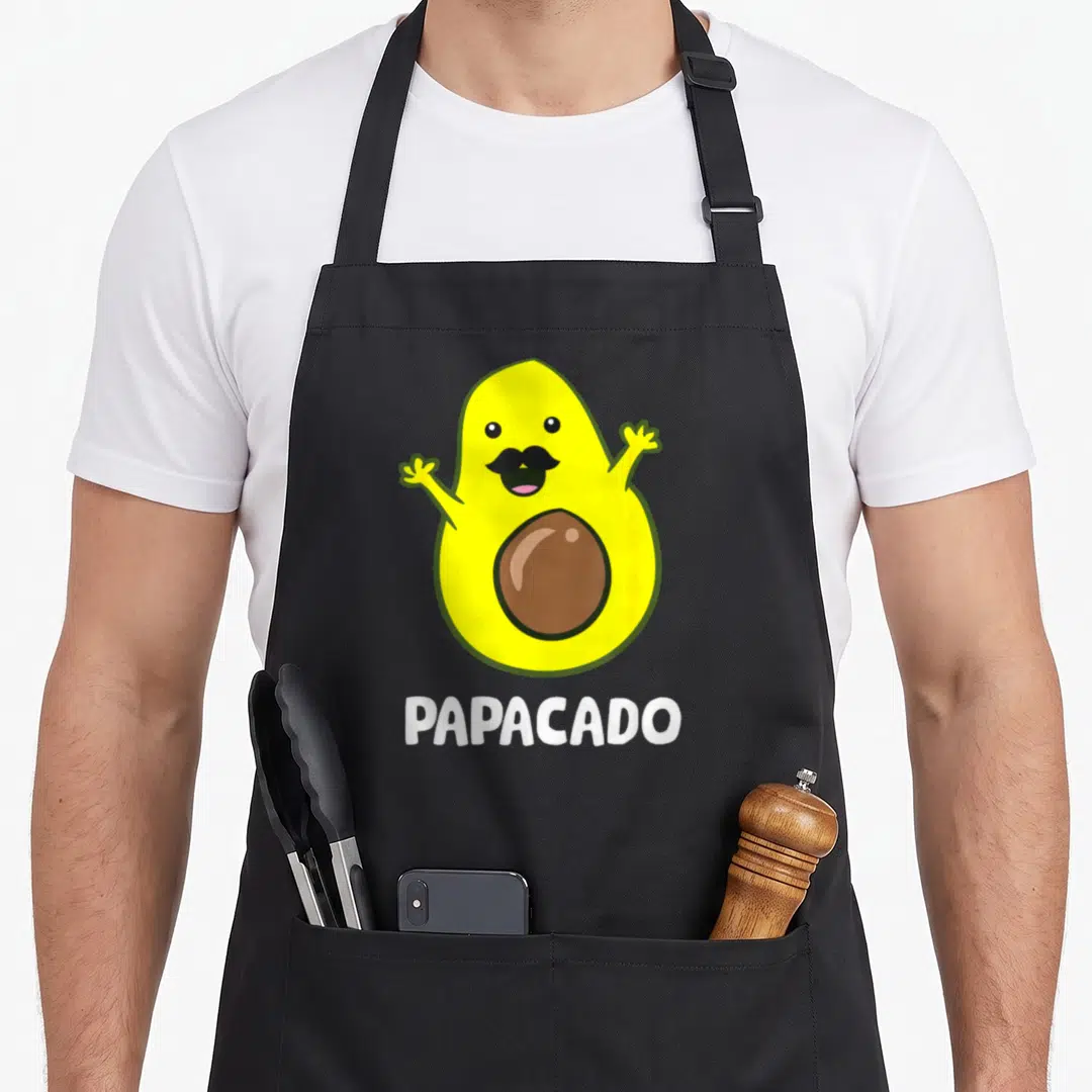 Vegan avocado apron - Yellow cartoon avocado with black mustache and brown pit above white PAPACADO text — shown on a black cotton canvas apron against a white background