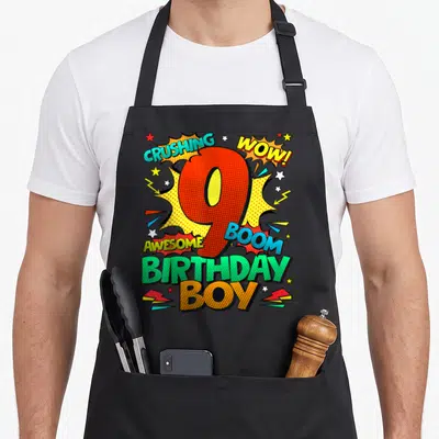 9th comic birthday apron - Red number 9 with CRUSHING WOW AWESOME BOOM BIRTHDAY BOY text in retro comic style — shown on a black cotton canvas apron against a white background