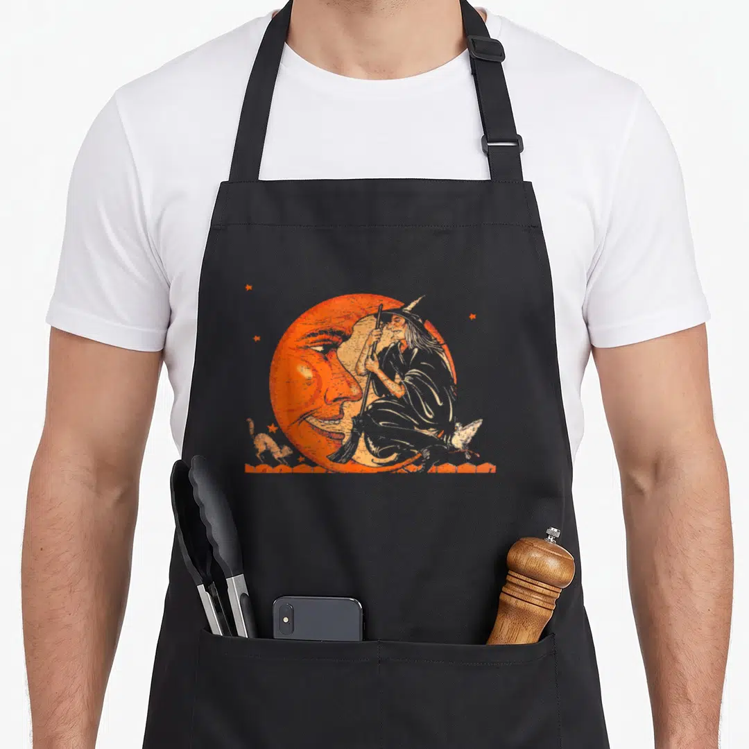 Moon halloween witch apron - Vintage line-art witch on crescent moon with broom and stars in orange and beige — shown on a black cotton canvas apron against a white background