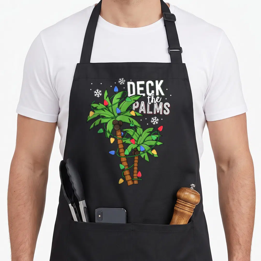 Deck christmas lights apron - Two palm trees wrapped in red blue and green lights with DECK the PALMS text — shown on a black cotton canvas apron against a white background