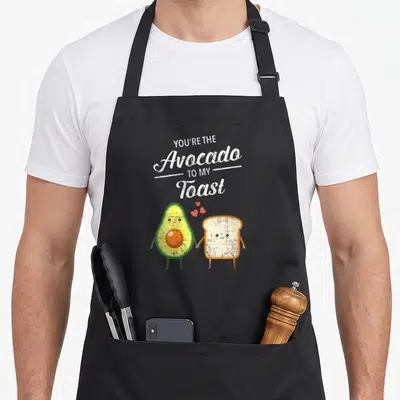 Couple toast apron - Cartoon avocado and toast holding hands with You're The Avocado To My Toast text and red hearts — shown on a black cotton canvas apron against a white background