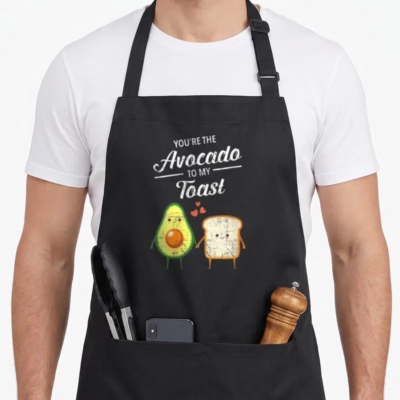 Couple toast apron - Cartoon avocado and toast holding hands with You're The Avocado To My Toast text and red hearts — shown on a black cotton canvas apron against a white background
