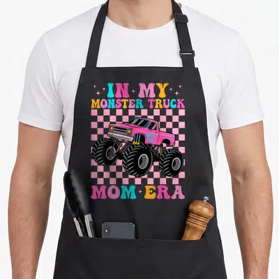 Era truck apron - Pink monster truck on checkered background with retro IN MY MONSTER TRUCK MOM ERA text — shown on a black cotton canvas apron against a white background