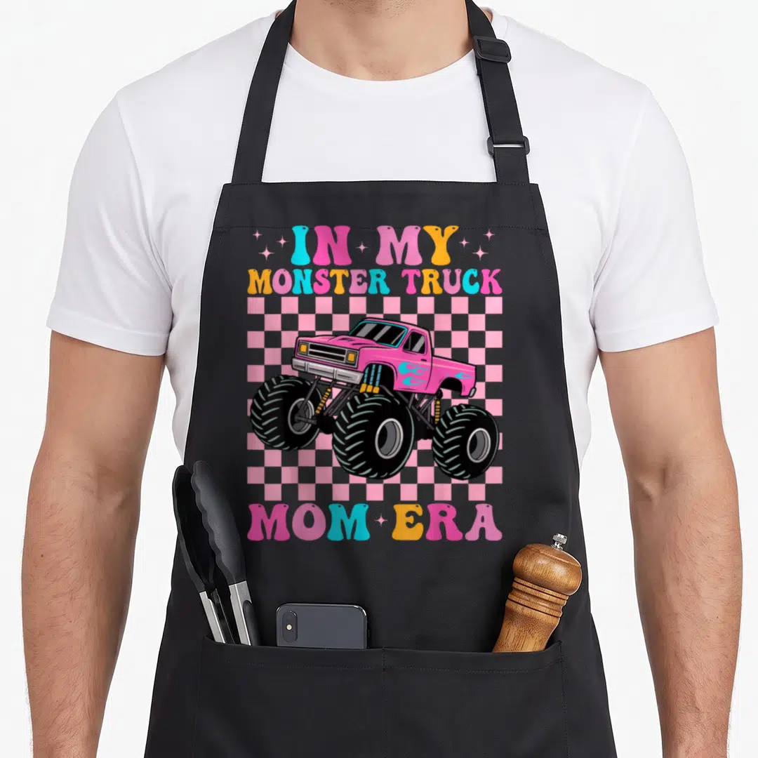 Era truck apron - Pink monster truck on checkered background with retro IN MY MONSTER TRUCK MOM ERA text — shown on a black cotton canvas apron against a white background