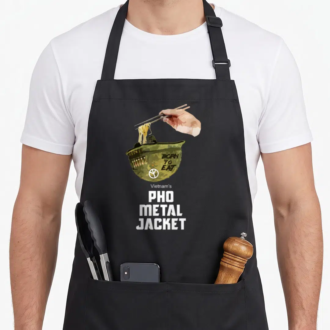 Metal pho apron - Cartoon olive helmet bowl with chopsticks lifting noodles and born to eat pho metal jacket text — shown on a black cotton canvas apron against a white background