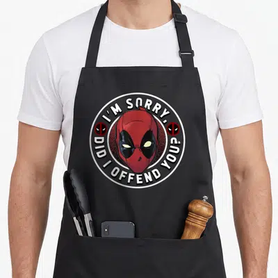 Offend sorry apron - Red Deadpool head surrounded by circular white text and two small mask icons on black background — shown on a black cotton canvas apron against a white background