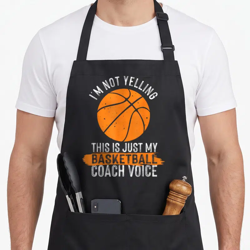 Basketball trainer dad apron - Orange basketball with white distressed text reading I'm not yelling this is just my — shown on a black cotton canvas apron against a white background