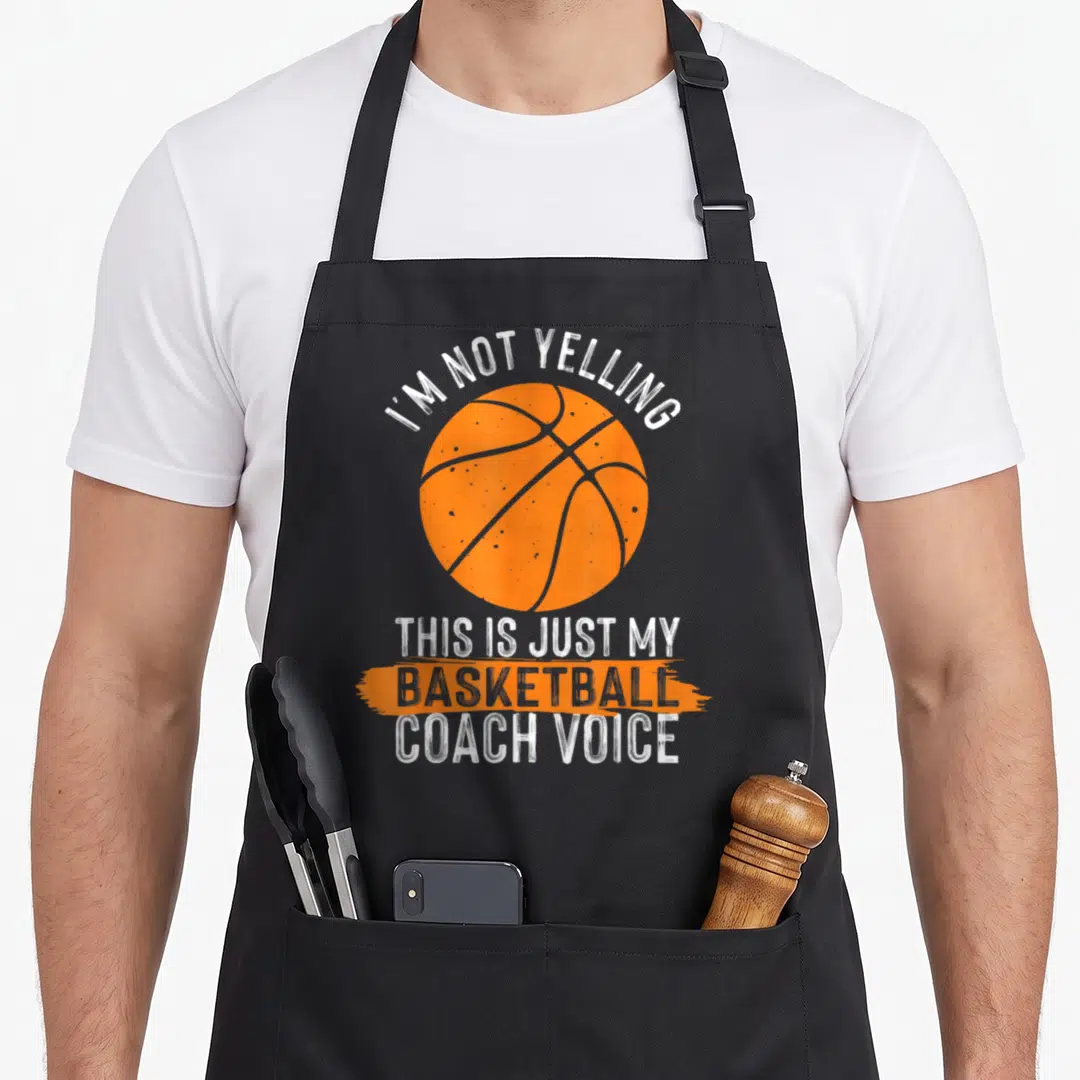 Basketball trainer dad apron - Orange basketball with white distressed text reading I'm not yelling this is just my — shown on a black cotton canvas apron against a white background