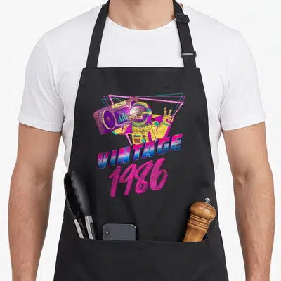 Born day 40th apron - Astronaut holding boombox with neon triangles above VINTAGE 1986 text in pink and cyan — shown on a black cotton canvas apron against a white background