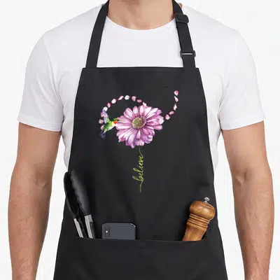 Believe hummingbird bird apron - Watercolor purple flower with green hummingbird and vertical believe script — shown on a black cotton canvas apron against a white background