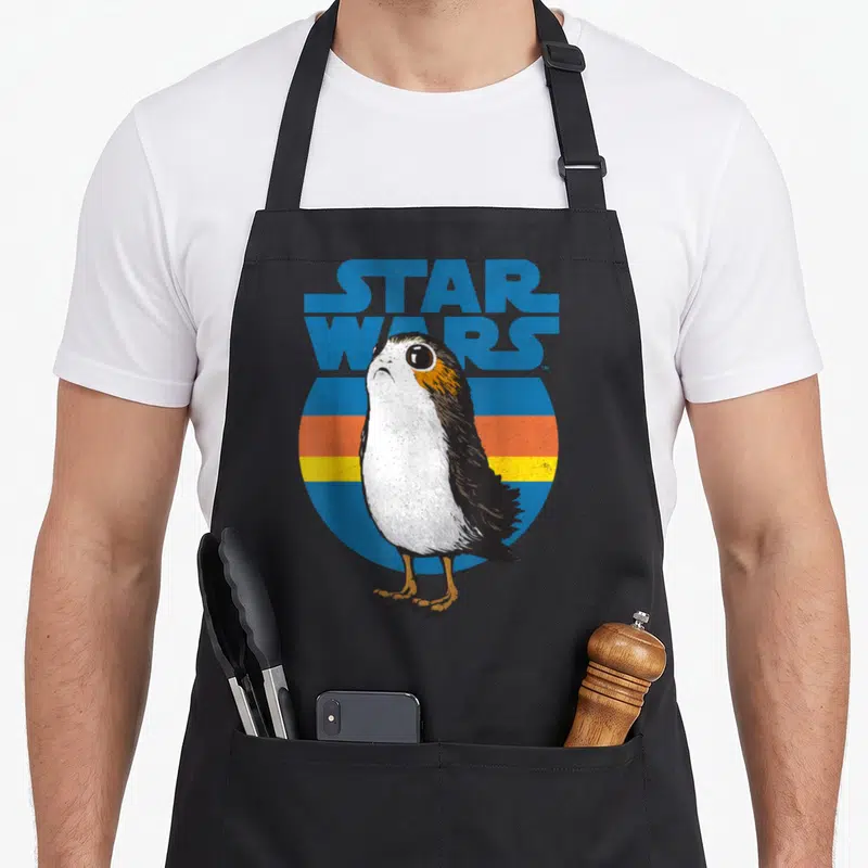 Porg stripe apron - Realistic porg bird in front of blue orange and brown stripes with Star Wars text — shown on a black cotton canvas apron against a white background
