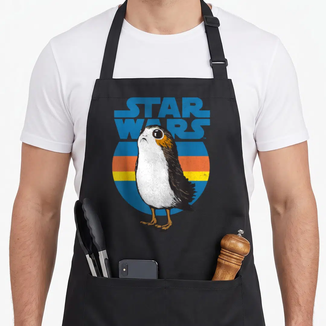 Porg stripe apron - Realistic porg bird in front of blue orange and brown stripes with Star Wars text — shown on a black cotton canvas apron against a white background