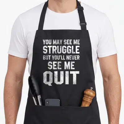 Workout sarcastic apron - White stacked text reading YOU MAY SEE ME STRUGGLE BUT YOU'LL NEVER SEE ME QUIT on black — shown on a black cotton canvas apron against a white background