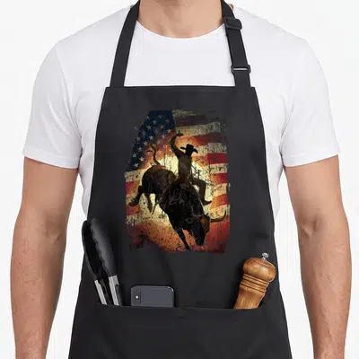 Rider cowboy apron - Bull rider silhouette on distressed American flag with red white blue yellow and black — shown on a black cotton canvas apron against a white background