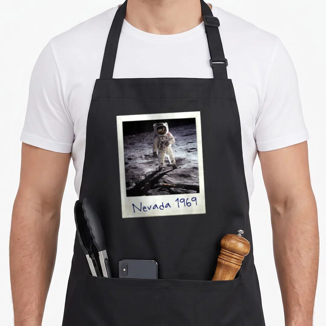 Fake patch apron - Polaroid photo of astronaut with handwritten Nevada 1969 text below — shown on a black cotton canvas apron against a white background