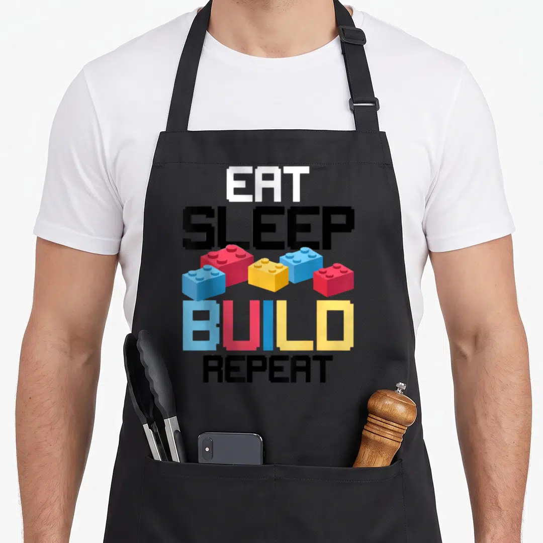 Eat building apron - Pixel text EAT SLEEP BUILD REPEAT with five red blue yellow white and black blocks — shown on a black cotton canvas apron against a white background