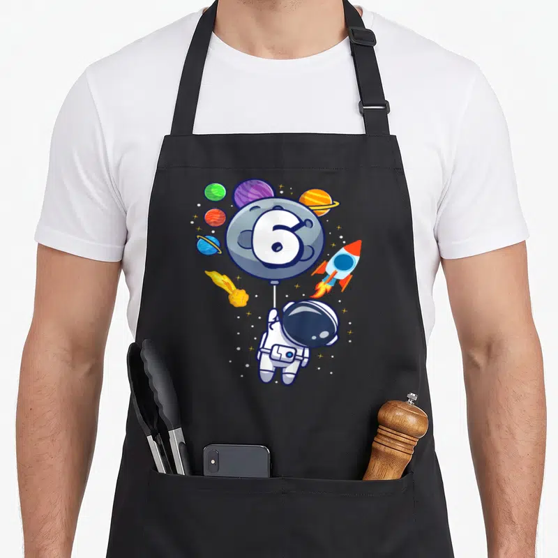 6th astronaut birthday apron - Cartoon astronaut holding grey balloon with number 6 surrounded by planets and rocket — shown on a black cotton canvas apron against a white background