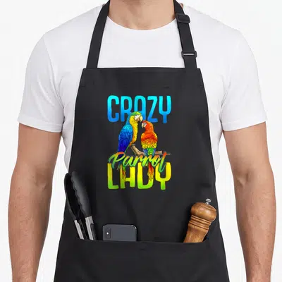Scarlett bird apron - Two parrots perched on a branch surrounded by CRAZY Parrot LADY text in blue and yellow — shown on a black cotton canvas apron against a white background