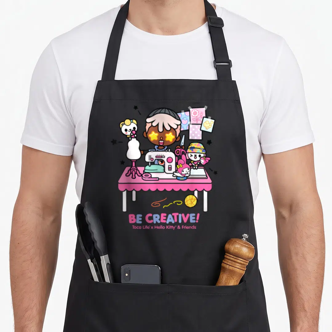 Toca creative apron - Cartoon person sewing at pink table with Hello Kitty characters and BE CREATIVE text — shown on a black cotton canvas apron against a white background