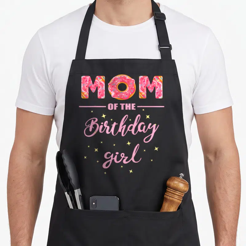 Birthday donut apron - MOM with pink donut O, Birthday girl script, yellow stars — shown on a black cotton canvas apron against a white background