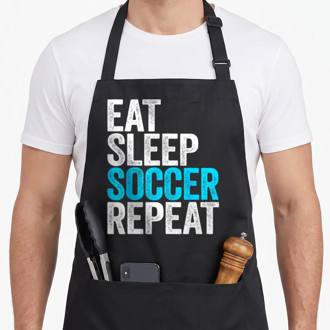 Eat soccer fan apron - Stacked distressed text reading Eat Sleep Soccer Repeat in white and light blue — shown on a black cotton canvas apron against a white background
