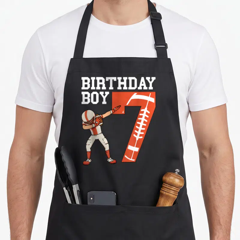 Football player birthday apron - Cartoon football player dabbing next to orange number 7 with BIRTHDAY BOY text — shown on a black cotton canvas apron against a white background