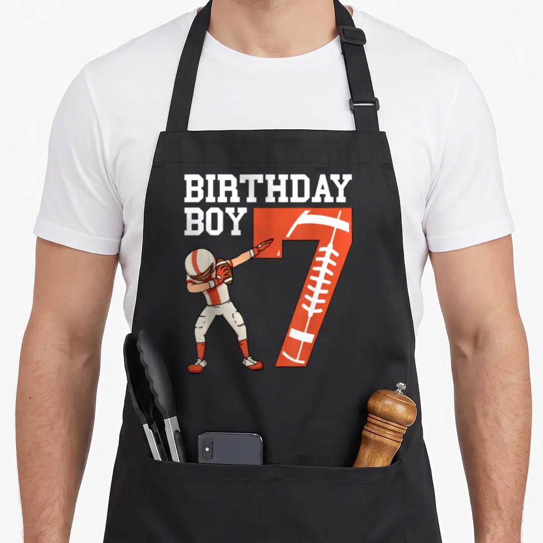 Football player birthday apron - Cartoon football player dabbing next to orange number 7 with BIRTHDAY BOY text — shown on a black cotton canvas apron against a white background