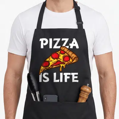 Cute food pizza apron - Cartoon pizza slice with PIZZA IS LIFE text in white yellow red and brown — shown on a black cotton canvas apron against a white background