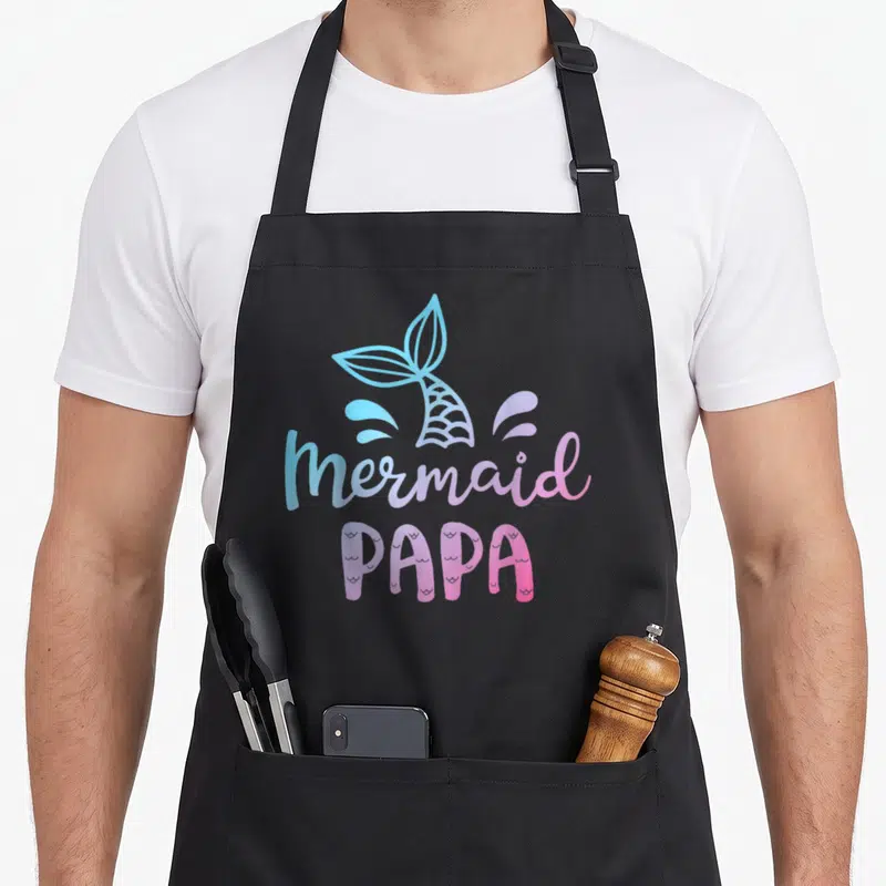 Merman grill daddy apron - Cartoon mermaid tail in blue and pink with text mermaid PAPA — shown on a black cotton canvas apron against a white background
