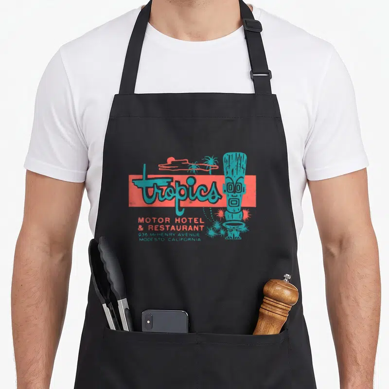 Tropic 50s style apron - Teal tiki statue next to coral rectangle with tropics text and Modesto California address — shown on a black cotton canvas apron against a white background