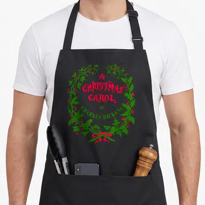 Wreath christmas apron - A Christmas Carol by Charles Dickens text inside a green holly wreath with red berries and bow — shown on a black cotton canvas apron against a white background