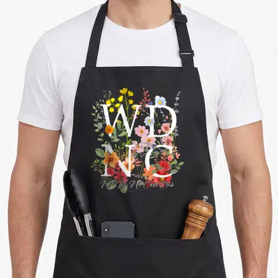 Wdnc floral apron - Stacked white WD NC letters over colorful watercolor flowers and We Do Not Care Club script — shown on a black cotton canvas apron against a white background