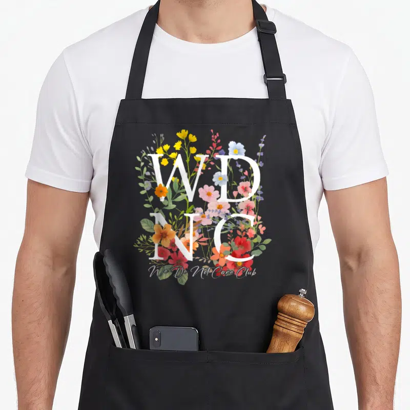 Wdnc floral apron - Stacked white WD NC letters over colorful watercolor flowers and We Do Not Care Club script — shown on a black cotton canvas apron against a white background