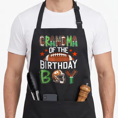 Game grandma aprons - Grandma of the Birthday Boy text with football and helmet graphic in brown and green — shown on a black cotton canvas apron against a white background